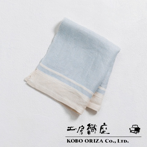 一條淺藍色純亞麻斜紋廚房拭巾，帶有米白色與淺藍色條紋邊飾，質地細緻，放在白色紋理背景上。