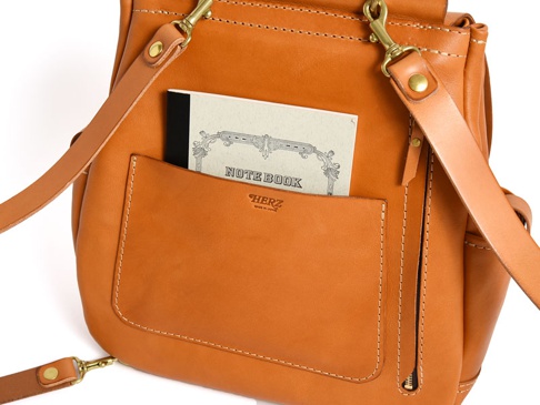 經典雙扣後背包，採用天然皮革製作，呈現溫潤的焦糖棕色。包款正面有一個開放式口袋，內含一本寫著「NOTEBOOK」的筆記本。包身縫線精緻，搭配黃銅色金屬扣環，展現職人手作質感。