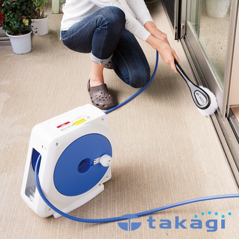 白色塑膠捲軸式灑水器，配有藍色圓形盤面和藍色軟管，軟管末端連接一個帶有白色刷毛的清潔刷，正在被使用清潔玻璃門。