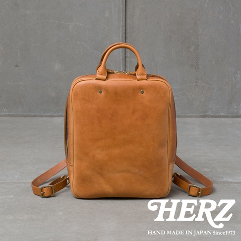 一個淺棕色皮革製成的方形後背包，配有頂部提把和可調節的肩帶，放在灰色水泥背景前。