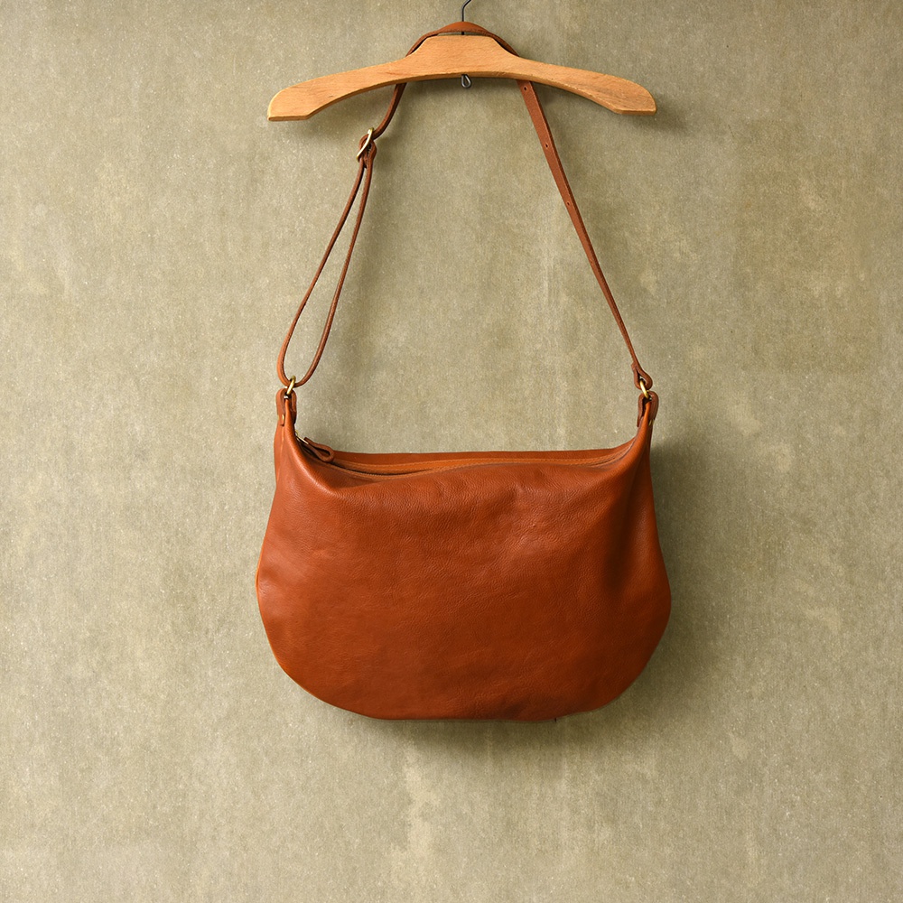 職人手作皮革Organ單肩半月包 G-15 M，採用棕色天然皮革製成，包身呈現經典半月形狀，搭配可調節的細皮革肩帶，以木製衣架懸掛在淺棕色紋理牆面上。
