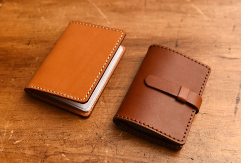 兩款皮革卡片收納夾，一款為淺棕色，另一款為深棕色，皆採用手工縫線，深棕色款式帶有皮扣設計，擺放於木紋背景上。