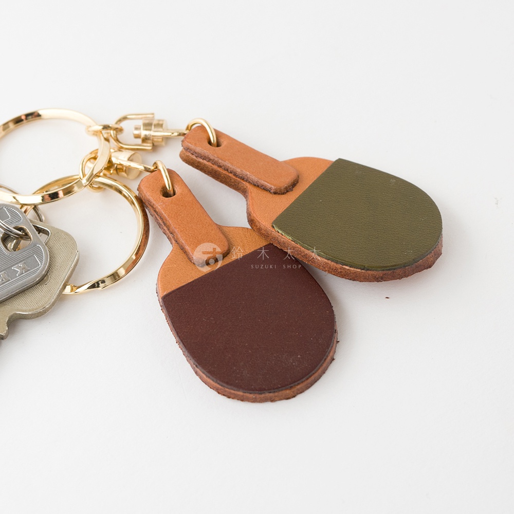 職人手作皮革桌球拍造型鑰匙圈，採用兩種顏色皮革拼接設計，一款為棕色與綠色，另一款為棕色與深棕色，搭配金色鑰匙圈。