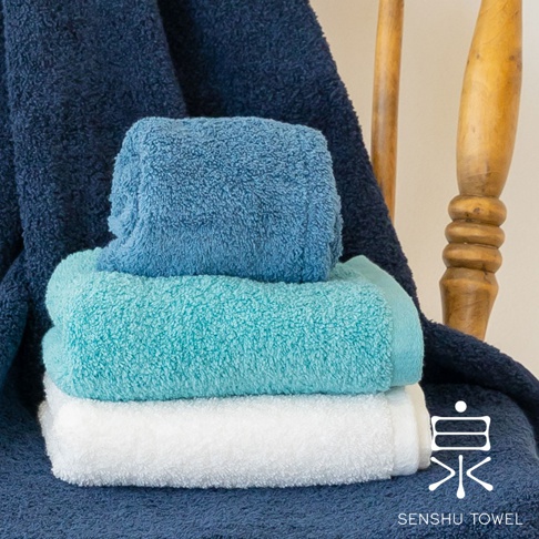 三條疊放的 envelopper 迷你浴巾，顏色由上至下為藍色、淺藍綠色和白色，材質皆為柔軟長纖維。