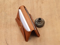 職人手作的雙開式卡片收納夾，採用棕色皮革材質，設有金色按扣，內部可收納白色卡片，旁邊擺放一個復古金屬旋鈕。