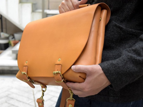 職人手作皮革雙扣兩用側背包，採用天然棕色皮革，設有兩個黃銅色金屬扣環和可調節的皮革肩帶，展現簡約復古風格。