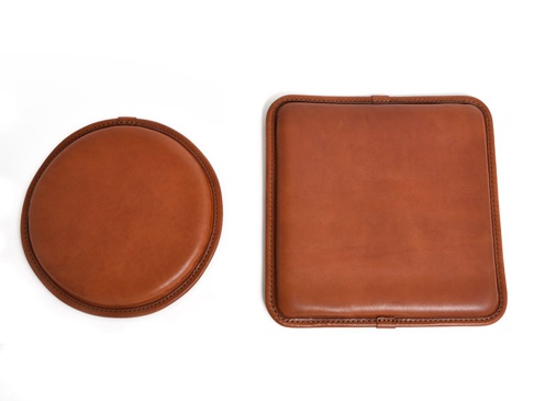兩個職人手作皮革坐墊，一個圓形，一個方形，均為暖棕色皮革材質，邊緣有精緻縫線裝飾，風格簡約。