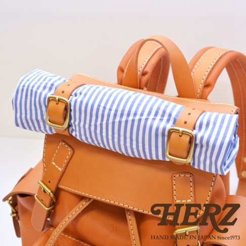 職人手作皮革復古風旅行後背包，以天然皮革製成，呈現經典棕色。背包頂部捲起處包裹著藍白條紋布料，並以皮革束帶及復古金色釦環固定，展現精緻工藝。