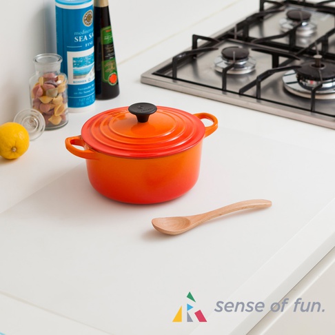 白色防滑料理台保護墊，墊上擺放著一個橘紅色圓形鑄鐵鍋，鍋蓋中央有黑色提把，鍋旁有一支木製湯匙，背景為廚房爐灶與調味品。