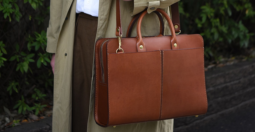 棕色皮革雙口袋兩用商務公事包，配有頂部提把和可拆卸肩帶，整體風格簡潔大方。