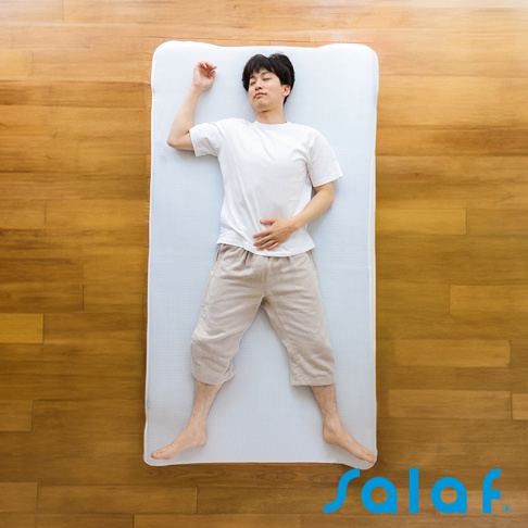 一名男性穿著白色T恤與淺卡其色短褲，仰臥在白色單人睡墊上，睡墊置於木紋地板上。