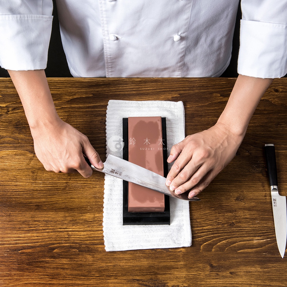 廚師正在一塊紅色磨刀石上打磨一把日本刀，旁邊還有一把未使用的刀。