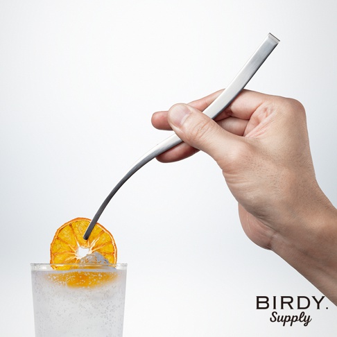 一只手使用銀色金屬製成的細長調酒夾，夾著一片乾燥的橘子片，準備放入裝有氣泡飲料和冰塊的玻璃杯中。