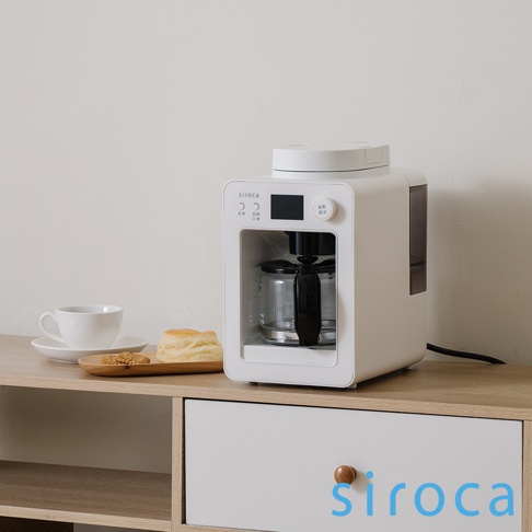 白色 Siroca SC-A3520 自動研磨咖啡機，配有玻璃咖啡壺、黑色手柄，頂部有白色蓋子。咖啡機正面有黑色顯示螢幕和按鈕，左側有一組白色杯子和木質托盤，上面放著司康餅和餅乾。