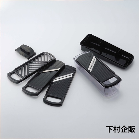 這是一款下村企販品牌的多功能刨絲器組合，包含一個透明收納盒、一個黑色蓋子以及三個可更換的刨絲刀片組件，各組件呈現黑色塑料材質與不鏽鋼刀片，整體以俯視角度展現，白色背景突顯商品結構與細節。