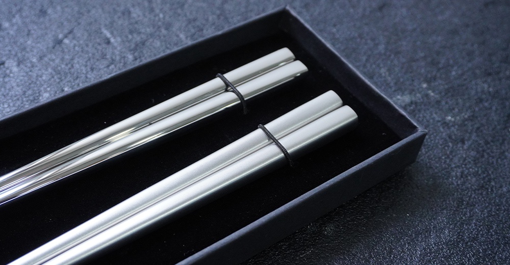 燕三条不鏽鋼筷兩雙，銀色，圓柱形，頂部有黑色細繩綁紮，放置於黑色紙盒內。