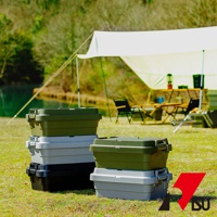 草地上堆疊著三款 TRUNK CARGO 可堆疊多功能耐重收納箱，分別為綠色、灰色與黑色。收納箱為塑膠材質，帶有銀色金屬扣環。背景是露營場景，有天幕、桌椅和湖泊。