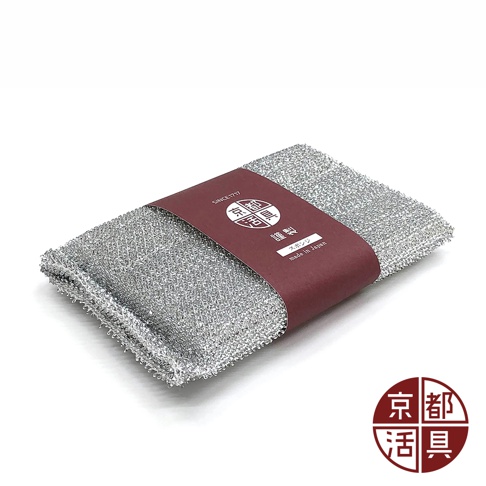 一個橫放的長方形菜瓜布，表面覆蓋著細緻的銀色金屬微粒，中心圍繞著酒紅色的紙質包裝帶，帶上印有京都活具的圓形標誌、日文產品說明及金色字體的 made in Japan 字樣，圖片右下角同樣附有該品牌的紅色圓形標誌，整體呈現簡約的廚房清潔用品質感。