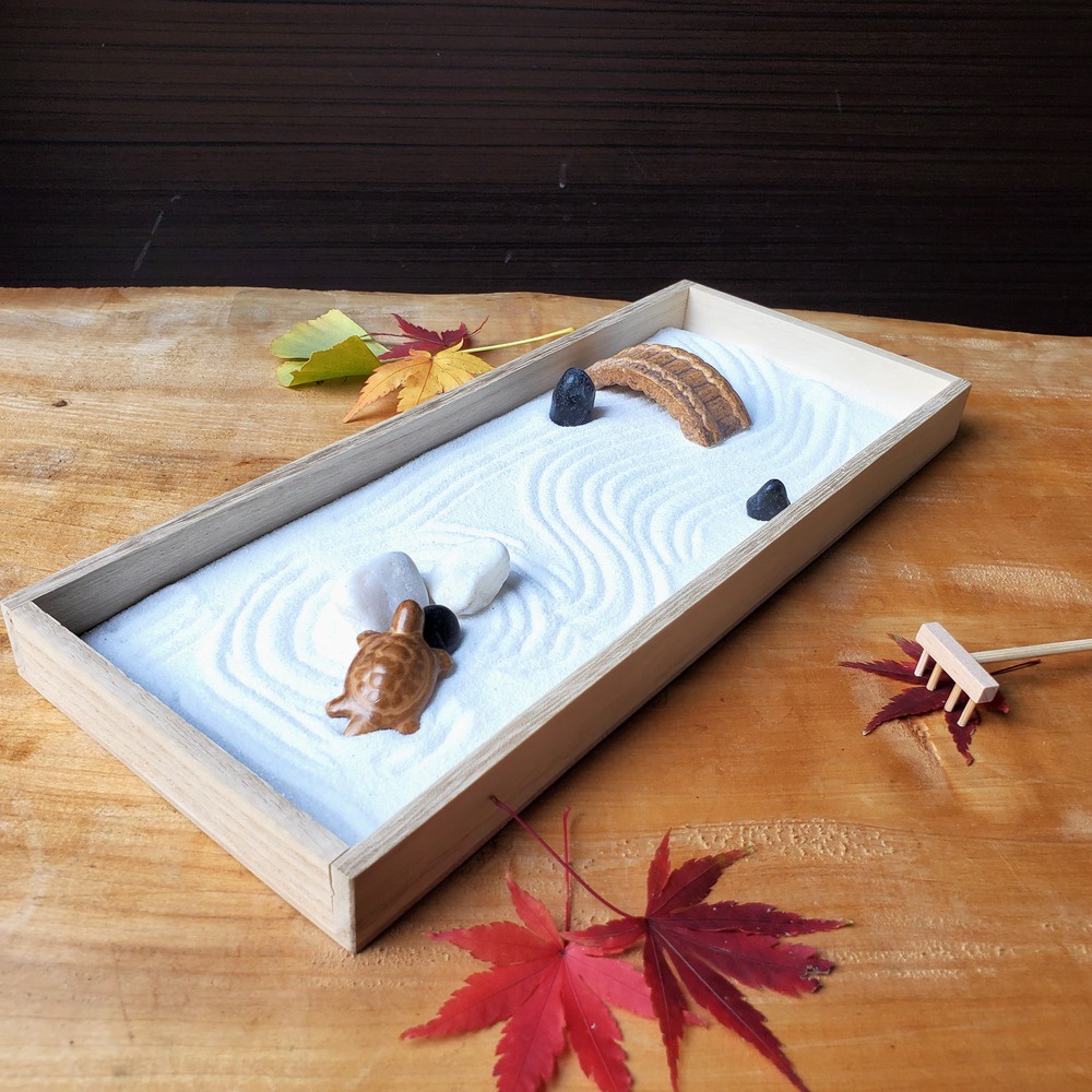 木製長方形盆中，鋪滿白色細沙，並以耙子畫出水紋。盆內有淺褐色陶瓷烏龜、白色鵝卵石、黑色小石塊，以及一座褐色木紋小橋。一旁有迷你木耙和紅色楓葉裝飾。