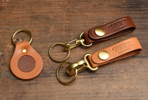 三款職人手作皮革鑰匙圈，包含一個棕褐色水滴形狀的鑰匙圈，中央為深棕色皮革圓片，以及兩個長條形皮革鑰匙圈，分別為紅棕色和淺棕色，皆附有黃銅色的金屬扣環和鑰匙圈。