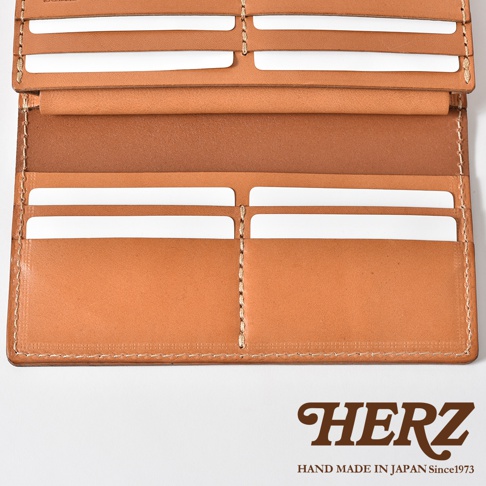 日本職人品牌 HERZ 手作皮革長夾，展開後的內部呈現，採用天然植鞣皮革製成，表面呈現溫潤的淺棕色澤。長夾內部配置多個對稱式卡片夾層，線條簡潔洗鍊，車縫工藝細緻，整體展現高品質皮革質感，下方印有品牌標誌與手工製造資訊。