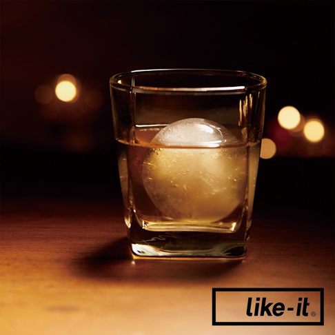 玻璃杯中盛有琥珀色液體，杯內有一個透明的圓形冰球。杯子置於木製檯面上，背景是模糊的暖色燈光。