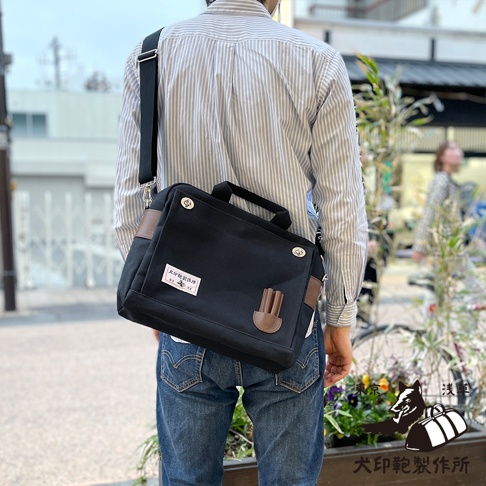 黑色純綿帆布製成，帶有皮革飾邊的橫式側背包，可肩背或手提，正面有品牌標籤。