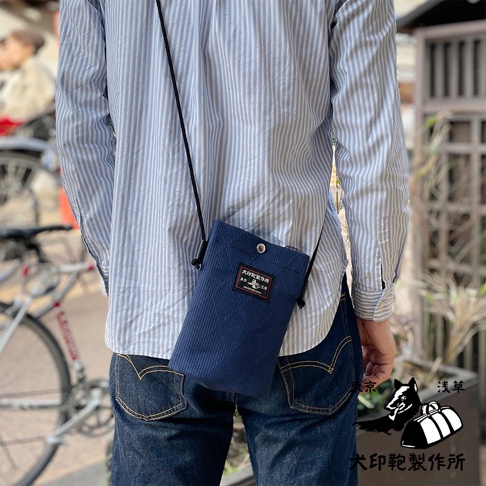 深藍色燈芯絨刺子織手機包，側面有品牌標籤，以斜背包方式掛於肩部。