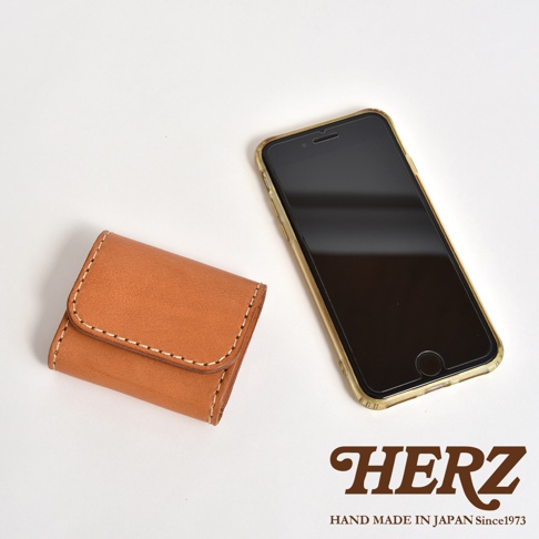 白色背景上放著一個日本HERZ手工製作的棕色皮革雙折零錢包，其表面有明顯的縫線細節，旁邊擺放了一支裝有透明保護殼的智慧型手機，右下角印有HERZ品牌標誌及相關文字。