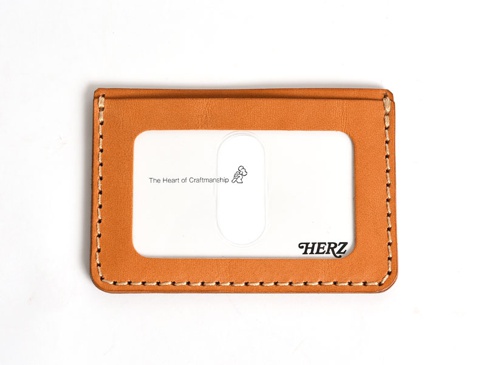 職人手作皮革橫式票券收納夾，採用天然鞣製皮革，呈現溫潤的橘棕色調，邊緣以米白色縫線工整車縫。正面中央設有透明PVC票券夾層，內嵌「The Heart of Craftsmanship」字樣與小人圖標，右下角壓印黑色「HERZ」品牌標誌。