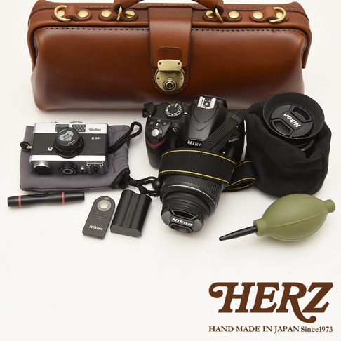 HERZ 品牌標誌與「HAND MADE IN JAPAN Since1973」文字，周圍擺放著皮革包、相機、鏡頭、電池及清潔工具。