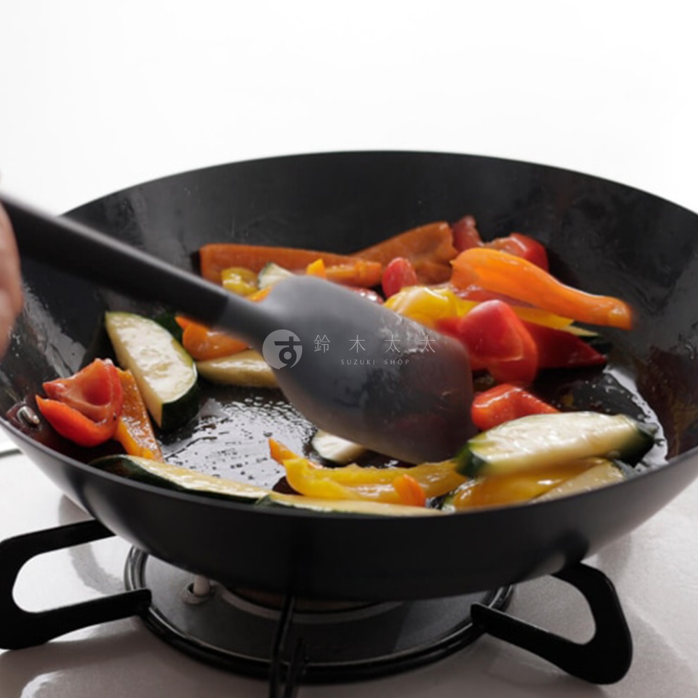 黑色鍋中正在烹炒切塊的蔬菜，包含紅蘿蔔、甜椒、節瓜及番茄，一只灰色矽膠鍋鏟正在攪拌，鍋鏟上有日文及 SUZUKI SHOP 字樣。