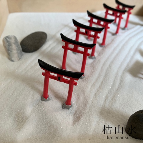 一排紅黑色鳥居模型，立於細白沙地上，旁邊散落著幾顆灰黑色石塊。