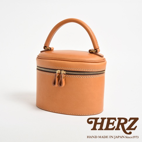 職人手作皮革圓筒型化妝手提包，以天然植鞣皮革製成，呈現溫潤的淺棕色。包款設有雙拉鍊開口，配有皮革拉環，頂部有圓弧形提把，整體造型簡潔且實用。