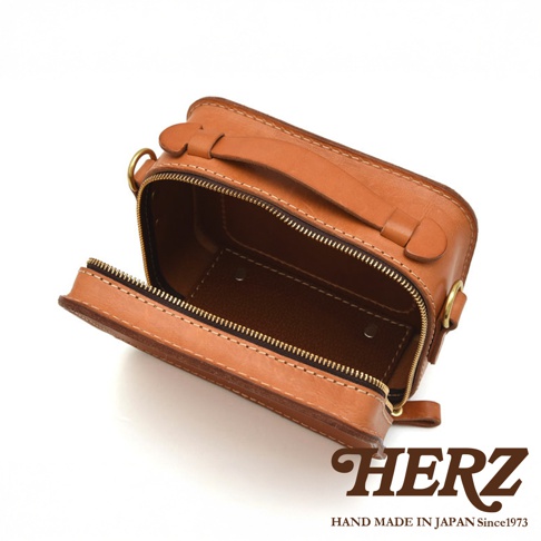 HERZ 品牌標誌，下方為 HAND MADE IN JAPAN Since1973 字樣。圖片呈現一只打開拉鍊的棕色皮革小方包，包包內部皮革顏色較深，拉鍊為金色。