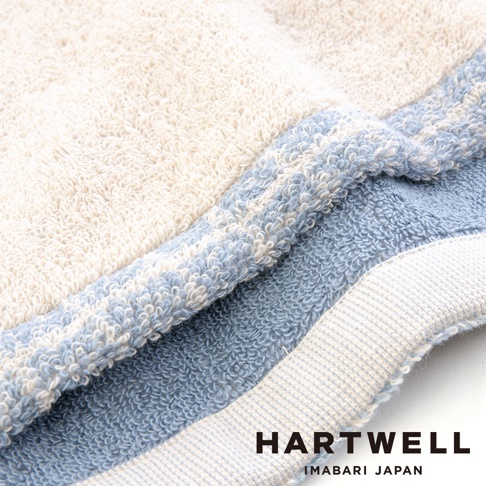 這張照片呈現了 Hartwell 今治午睡貓咪雙色浴巾的細節，特寫展現了毛巾細膩柔軟的棉質毛圈質感，米色與柔和藍色交織的雙色設計呈現出舒適的層次感，右下角印有 Hartwell Imabari Japan 品牌字樣。