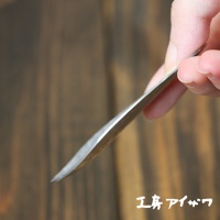 一隻手拿著一把銀色不鏽鋼製的茶勺，勺身呈現細長的彎曲狀，邊緣光滑，背景為木紋。