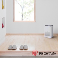 白色簡約風格情境式空氣清淨機 IAP-A25，方形機身，頂部有控制面板，前側有網狀進氣孔，底部有藍色指示燈，放置於木質地板上，背景有窗戶及綠色植物。