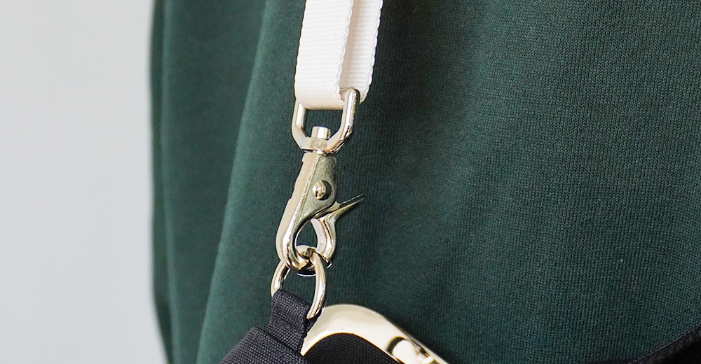 圖片上的斜背包掛帶，採用白色的帆布材質，搭配銀色的金屬扣環和鉤子，掛在深綠色的衣物上。
