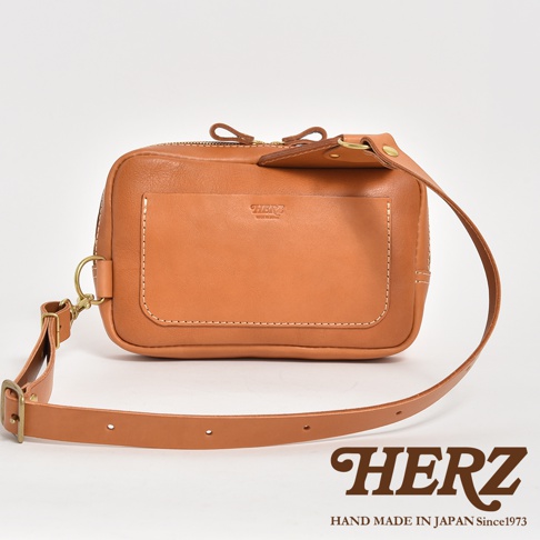 職人手作皮革雙口袋斜背包，主體為棕褐色皮革，設計簡約，前方有一明顯的方形貼袋，袋上有壓印品牌標誌。包包兩側有金屬D型環，搭配可調節的皮革斜背帶。