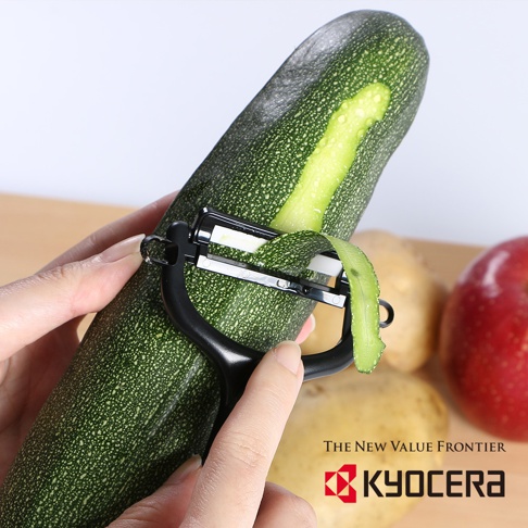 黑色陶瓷削皮刀正在削綠色櫛瓜的皮，旁邊有馬鈴薯和蘋果。