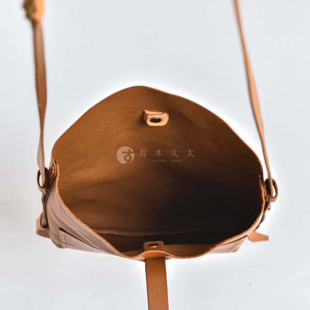 由上方俯瞰視角拍攝的日本HERZ職人手作皮革單肩包，棕色牛皮材質呈現柔軟細緻的紋理。包包內部空間開闊，中央標有鈴木太太的品牌字樣，開口處設有金屬扣環與皮帶翻蓋，兩側連接細長肩帶，整體造型簡約俐落。