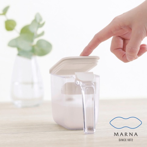 一隻手正輕壓 MARNA 密封調味料盒的蓋子，該調味料盒呈現透明質感並設有白色手把，盒內裝有白色粉末狀物品，背景為乾淨的淺色空間並擺放一盆綠色植物。