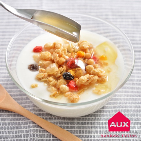 不鏽鋼挖蜂蜜匙正在將金黃色蜂蜜淋入裝有優格、麥片和乾燥水果（如草莓、藍莓和蔓越莓）的透明玻璃碗中。