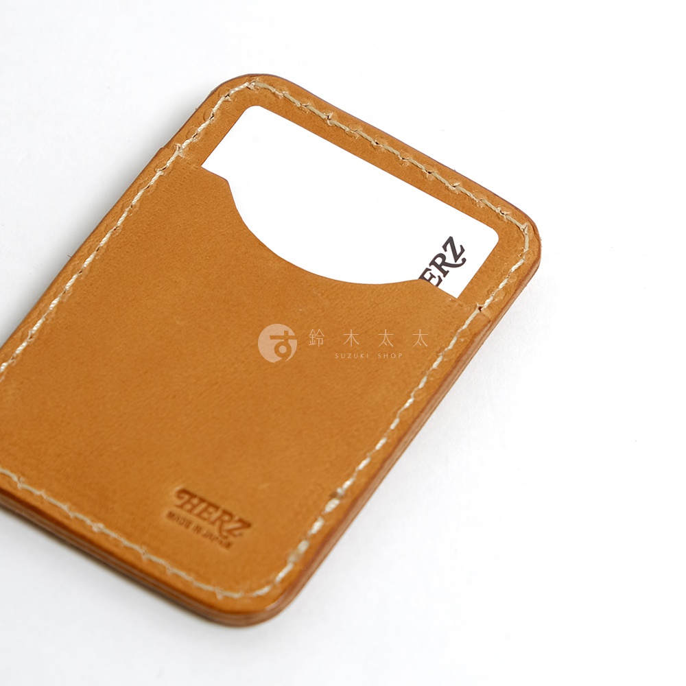 職人手作皮革名片夾，淺棕色皮革，手工縫線，右側有一個插入名片的開口，左下方壓印有HERZ品牌標誌和MADE IN JAPAN字樣。
