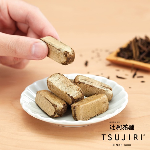 一隻手捏著一塊長條狀的辻利茶舖精選茶酥，下方白色波浪邊碟子裝著四塊同樣的茶酥，背景為木質桌面，右上方散落著少量茶葉與木製容器，畫面呈現溫潤的茶點質感。