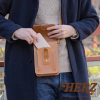 職人手作皮革休閒散步包，主體為淺棕色天然皮革，可見細膩縫線與金屬釦，包款設計可放置手機，適合日常攜帶。