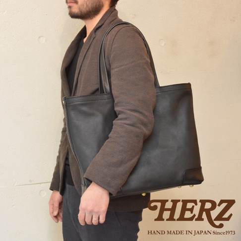 一個男人側身肩背著一個黑色皮革托特包，包包底部有金色鉚釘裝飾。