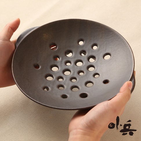 一隻手拿著深棕色、圓形的美濃燒多用蒸盤，盤面佈滿大小不一的透氣孔，盤邊有兩側把手。