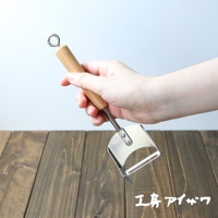 一隻手正握著 VEGET 不鏽鋼木柄削皮刀，削皮刀具有光滑的木質手柄和銀色的不鏽鋼刀頭，刀頭設計為弧形，刀刃鋒利，適合削皮。